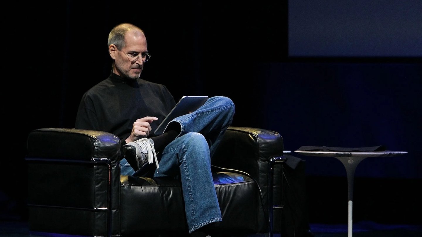 Steve Jobs sentado en un sofá con las piernas cruzadas y un iPad en las manos